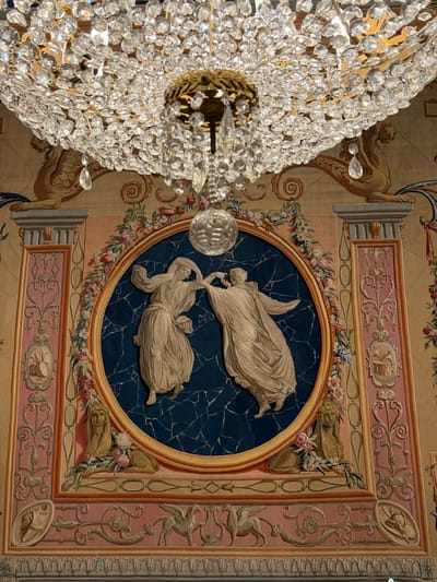 The image shows a close-up of a crystal chandelier hanging above a tapestry. The tapestry depicts two figures in flowing robes dancing within a circular frame, surrounded by floral motifs and architectural elements. The figures appear to be engaged in a graceful movement, possibly a duet. The overall scene is rich in detail and suggests an opulent setting. This particular tapestry is located in the Royal Tapestry Hall of the Monastery of San Lorenzo de El Escorial, a historical residence of the King of Spain, situated in San Lorenzo de El Escorial, Spain.