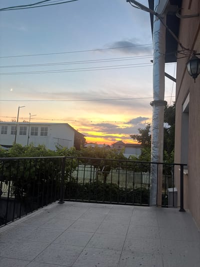 The image depicts a sunset viewed from a balcony in the city of Dolni Dabnik, Bulgaria. The sky is filled with warm hues of orange, yellow, and pink, with scattered clouds. A crescent moon is visible in the upper left portion of the sky. In the background, there are several buildings, including a white structure with windows and a dark-colored roof. Trees and greenery surround the buildings. Electrical wires crisscross the sky. On the right side of the image, a portion of a building with a light brown exterior wall is visible, along with a black outdoor lamp. The balcony itself is tiled with square grey tiles and has a black metal railing. A large, silver-covered pipe runs vertically along the side of the building.