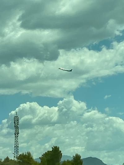 A distant, white airplane with a dark tail is captured in flight against a partly cloudy sky. The clouds are a mix of light gray and white, with patches of blue sky visible. In the foreground, a tall, metal telecommunications tower stands prominently on the left side of the frame. Below the tower and to the right, the tops of green trees are visible, suggesting a natural landscape. In the far background, the faint outline of a mountain range can be seen against the horizon. The image appears to be taken during the daytime under fair weather conditions.