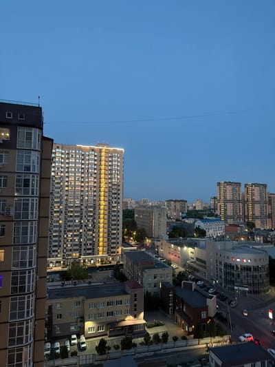 This media file presents an aerial view of a cityscape in Azov, Russia, during the twilight hours. The scene is dominated by numerous multi-story buildings, characteristic of urban development. The sky is a clear, deep blue, indicating the transition from day to night. Buildings of varying architectural styles are visible, with many windows illuminated, suggesting that people are inside and active. Some buildings are modern high-rises, while others appear older and more traditional. Roads and parking areas are interspersed throughout the urban landscape, with a few vehicles visible. Trees and other greenery are present, adding some natural elements to the built environment. The lighting conditions suggest it is either early evening or late evening, with artificial lights from the buildings and streetlights beginning to dominate the scene. There is no visible text in the image. The overall impression is one of a bustling, inhabited city as it winds down for the night.