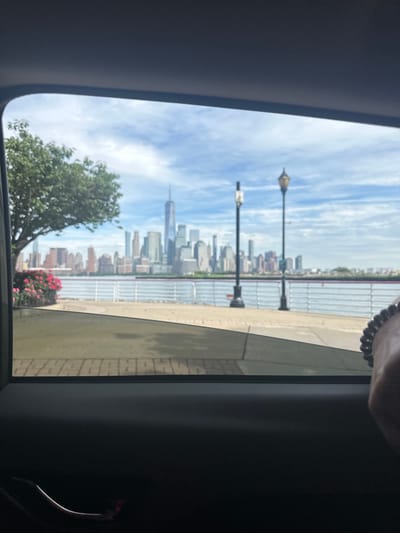 A view of the New York City skyline is framed by a car window, likely in Jersey City, New Jersey. The One World Trade Center spire is visible amongst the other skyscrapers. The foreground shows a tree and some flowers, as well as a sidewalk and lampposts. The sky is partly cloudy.