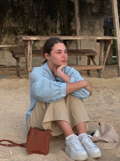 A young woman with brown hair pulled back sits on a sandy beach. She is wearing a light blue button-down shirt and tan pants. Her left arm is bent and resting on her right arm, with her hand supporting her chin as she looks to the right. In front of her are a small brown purse and a patterned tote bag. She is wearing white platform sneakers. Behind her is a weathered wooden structure with benches and a rough concrete wall. The ground is covered in sand, and a plastic water bottle is visible in the background. The location is the beach in Tong Yang, Thailand.