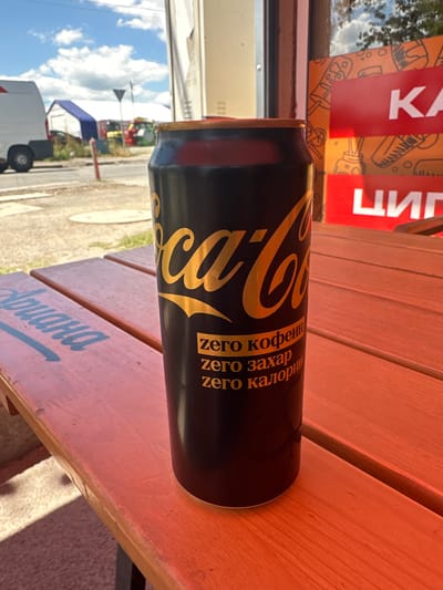 A close-up, eye-level shot features a black Coca-Cola can with gold lettering, positioned on a weathered orange wooden picnic table. The can displays the Coca-Cola logo and text in gold, which reads "zero кофеин", "zero захар", and "zero калории" in Cyrillic script. The background reveals an outdoor setting with a street and some buildings under a partly cloudy blue sky. In the distance, a white van is visible, along with other vehicles and structures, suggesting an urban or semi-urban environment. The overall lighting indicates it is daytime, likely sunny. This scene is located in Voluyak, Bulgaria.
