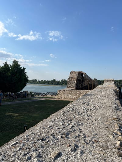 This media file captures a sunlit day at a historical site, likely a fortress or castle, located in Belene, Bulgaria. The image is oriented vertically, providing a sweeping view from a high vantage point.
The most prominent subject is a thick, ancient stone wall that dominates the right side of the frame. Its rough, weathered texture and irregular stone composition suggest a long history. To the left of the wall, a body of water, possibly a river or lake, stretches into the distance, bordered by a dense line of green trees on the opposite bank. The water is calm and reflects the clear blue sky.
In the foreground, a large, dark green conifer tree stands to the left, its branches full and lush. Below and to the right of the tree, a grassy area is visible, with a pathway running through it. A few people are scattered on the grass, some appearing to be seated, suggesting a leisurely visit or perhaps a small gathering. A wooden fence with a crisscross pattern runs along the edge of the grassy area near the water, and a dark metal structure, possibly part of the historical site's infrastructure or a viewing platform, is visible near the fence.
Further into the frame, a section of ruined stone structure is visible, partially supported by exposed wooden beams and scaffolding, indicating ongoing restoration or preservation efforts. The sky is a vibrant blue with scattered white, wispy clouds, suggesting fair weather. The strong sunlight casts shadows, implying it is likely late morning or early afternoon. The overall mood is serene and historical, with a sense of openness and natural beauty. No visible text is present in the image.