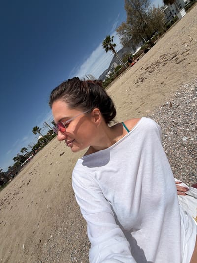 A woman wearing a white top and rose-tinted sunglasses is lying on a beach in Glifada, Greece. The sky is blue with some clouds. The beach is sandy and has small pebbles. In the background are buildings and palm trees. The woman is the primary subject of the image.