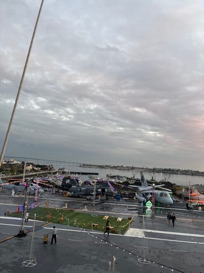 An aircraft carrier deck displays helicopters and airplanes. Some are decorated with holiday lights. People are walking on the deck. A strip of artificial turf is laid out for games. A skyline with buildings and a bridge is visible in the background. The sky is overcast and the image appears to be taken at dusk. A rope is visible in the foreground.