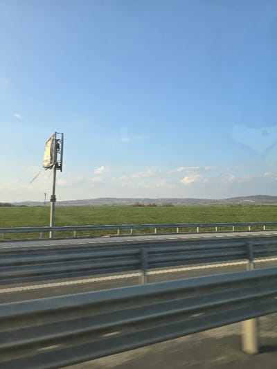 A photographic image captures a rural highway scene, consistent with the vicinity of Rupkite, Bulgaria, from the perspective of a moving vehicle. In the foreground, multiple parallel rows of corrugated metal guardrails line the edge of a multi-lane asphalt road, which displays white lane markings. Beyond the guardrails, a vast, flat expanse of vibrant green agricultural field extends towards the horizon under a clear, light blue sky featuring scattered white cumulus clouds near the distant tree line and rolling hills. To the left, a tall, slender metal pole rises from the green field, supporting an electronic signboard or communication equipment with a metallic frame and a partially damaged white panel, from which a strip of material hangs. No people, specific subjects, or direct actions are observable within the frame.