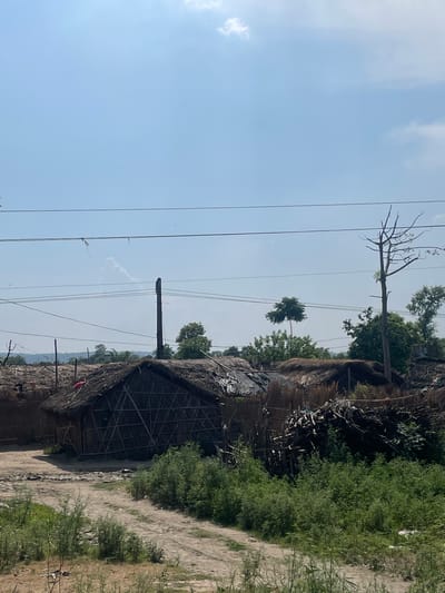 The image shows a landscape in Katauhar Kalan, India. A hut with a thatched roof stands in the foreground, and a dirt path runs along the right edge of the frame. In the background, there is a cluster of trees and vegetation. Overhead power lines stretch across the sky.