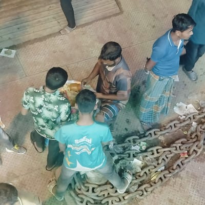The image captures an outdoor scene in Bhola, Bangladesh, on what appears to be a ferry deck or dock constructed with diamond-plate metal flooring. Large, heavy-gauge maritime chains are prominently laid across the right side and foreground, interspersed with discarded paper, plastic wrappers, and other debris.
Five adult males are discernible within the frame. At the center, a man wearing a brown and blue striped long-sleeved shirt and a patterned lungi is seated or squatting, actively engaged with items in a large, round metal tray. The tray contains various food products, possibly snacks or pastries, some wrapped in leaves. Two other men face this vendor: one, wearing a green floral short-sleeved shirt, stands to the vendor's left, while another, wearing a teal 'safal' branded t-shirt, squats on the chains in the foreground.
To the right, a man in a blue polo shirt with white collar trim and a plaid lungi stands with his hands behind his back. The legs and feet of a person in dark trousers and sandals are visible in the top-left, and part of a head and shirt appear in the bottom-left. All individuals are casually dressed.