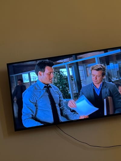 Two men are visible on a television screen. The man on the left, who is closer to the viewer, wears a blue dress shirt and a dark tie. He is looking down and appears to be speaking. The man on the right wears a gray suit jacket over a blue collared shirt and a dark vest. He is holding a light blue paper and looking at it. The scene appears to be set indoors, possibly an office, with windows in the background. The television is mounted on a wall painted a muted yellow-brown color. There are no visible elements that specifically indicate the location is Acones Dārzs, Latvia.