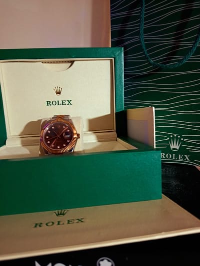 The image shows a close-up of a Rolex watch displayed in its green presentation box. The box is partially open, revealing the watch nestled inside on a cream-colored cushion. The watch itself has a gold bezel and bracelet, with a dark-colored face and diamond hour markers. Above the watch, the Rolex logo, a gold crown, is printed on the inside of the box lid, followed by the word "ROLEX" in green text.
In the background, a dark green shopping bag with a pattern of thin white wavy lines is visible. The bag also features the Rolex crown logo and the word "ROLEX" printed in white. The overall scene suggests a luxurious setting, likely a retail environment or a display of high-end goods. The lighting is somewhat dramatic, casting shadows and highlighting the metallic sheen of the watch and box. There are no people visible in the image. The visible text includes "ROLEX" and the Rolex crown logo, appearing multiple times on the box and bag. The scene is devoid of any specific environmental cues that would indicate time of day, weather, or a particular emotion. The context of "Ashkelon, Israel" is not directly observable from the visual information provided in the image itself.