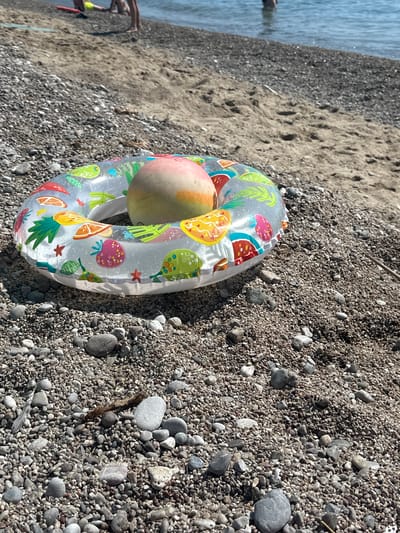 An inflatable swim ring lies on a pebble beach near the water. The swim ring is transparent with colorful fruit patterns, including strawberries, watermelons, and citrus slices. A light-colored fruit, possibly a peach or apricot, rests inside the swim ring. The beach is covered in small, grey and brown pebbles. In the background, the blue water of the sea is visible, with the faint figures of people wading in the shallows. The location is Đurovići, Montenegro.