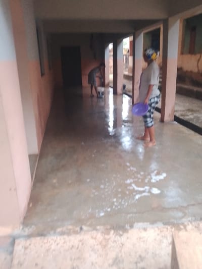 Two women are cleaning a wet, concrete floor under a covered walkway or corridor. One woman is bent over, appearing to scrub the floor with a bucket nearby. The other woman stands closer to the camera, holding a purple plate. Both women are barefoot. The setting is an open-air building with visible support pillars and doorways. The floor shows signs of soap and water use.