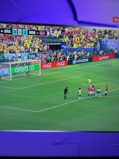 The image depicts a soccer match with a scoreboard indicating "BRA 0 - 2 NOR". The scoreboard also displays the FIFA World Cup logo. The field is green with white markings. In the foreground, several players are positioned, and one player in a yellow jersey is about to kick a soccer ball. A goalkeeper in green is near the goal. In the background, a large crowd of spectators is visible, many wearing yellow and red. Advertising banners for sponsors like Coca-Cola, Hyundai, and Lenovo are displayed around the stadium. The sky is not visible in the frame. The image does not contain any information or visual cues to suggest the location is Ognianovo, Bulgaria.