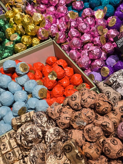 This is a close-up, top-down shot of a colorful assortment of individually wrapped chocolates, likely displayed in a store or market. The chocolates are arranged in bins or sections, each section filled with candies wrapped in distinct foil colors. From left to right, there are sections of teal, pink, purple, yellow, green, orange, light blue, and gold/brown. Many of the wrappers have visible text. Some wrappers, particularly those in the pink, yellow, and teal sections, feature a gold circular sticker with the text "LUX CHOCO" and Arabic script. Other wrappers, in the gold/brown and some of the purple sections, are adorned with intricate patterns and the text "La Fontaine" and "Lezatne". The overall impression is one of abundance and variety, with a focus on the visual appeal of the different colored wrappers and the details of the chocolate packaging. The lighting suggests an indoor setting, possibly a confectionery shop or a display at a food festival. There are no people or animals visible in the frame. The scene is static and focuses solely on the products. The text suggests these are premium or artisanal chocolates.