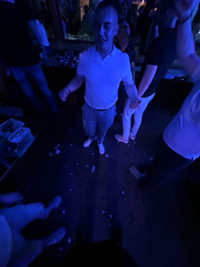 The image shows an interior space in Panagyurski kolonii, Bulgaria, with a group of people in low, blue lighting. One man is predominantly visible, wearing a white shirt and light-colored shorts, facing the camera with a smile. Other people are partially visible, with limbs and torsos entering the frame from different angles. There is debris, possibly confetti or torn paper, scattered on the floor. A serving tray with food containers is visible in the lower part of the frame. A window is on the left, partially showing an exterior view.