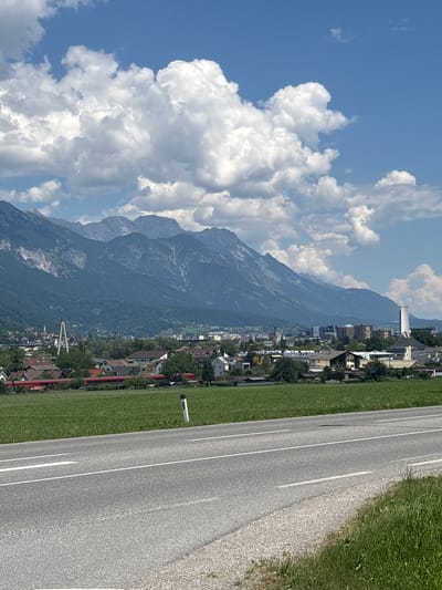 The image shows a wide, paved road in the foreground, with white lines indicating traffic lanes. Beyond the road is a vast, vibrant green field. In the middle ground, a town is nestled against a backdrop of imposing, lush green mountains. The town features a mix of residential buildings with red roofs and modern commercial structures. A tall, distinctive white building with a sharp, angled roof is prominent in the center-right of the town. A long, red train is visible traversing the landscape just behind the field. Above the town and mountains, a bright blue sky is dotted with fluffy white clouds. The scene appears to be a scenic view of Innsbruck, Austria, on a clear day.