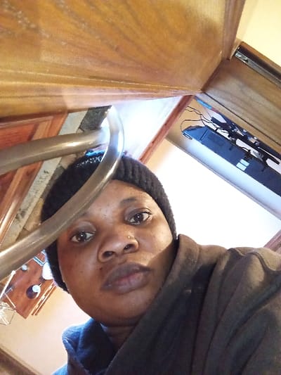 An adult female is depicted in a close-up, low-angle selfie, positioned indoors in Madinaty, Egypt. The subject, with dark skin, wears a black knit beanie and a dark-colored garment, possibly a jacket or top. A small stud piercing is visible on her right nostril. She maintains a neutral expression, looking directly at the camera. A curved, metallic railing extends diagonally across the upper left of the frame, partially obscuring her head. The background on the left features wooden paneling or furniture, while the right background shows a lighter wall or opening, with what appears to be a dark electronic device, possibly a soundbar or console, and associated cables resting on a surface below it. The environment is an interior space, likely domestic, with ambient lighting.