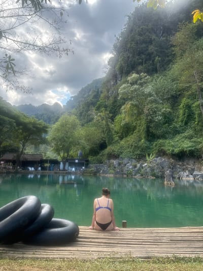 A woman in a swimsuit sits on a bamboo raft, facing a serene, green-tinted lake. Lush, green cliffs rise steeply, framing the scene under a cloudy sky. Several black inner tubes are stacked on the raft. In the distance, floating structures line the water's edge. The photograph captures a tranquil moment surrounded by natural beauty.