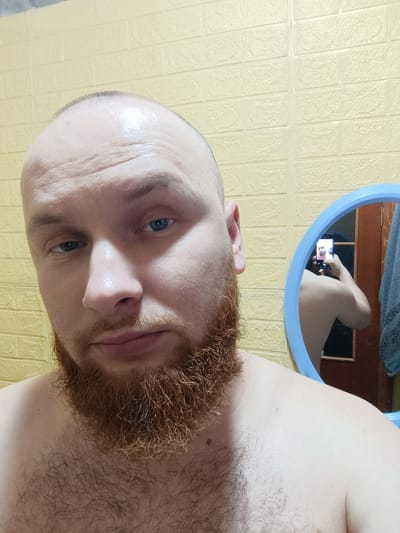 A man with a shaved head and a long, auburn beard takes a selfie in a bathroom. The man is shirtless, and his chest is hairy. Behind him, a yellow brick-patterned wall is visible. A blue-rimmed mirror reflects the man taking the photo, showing his back and shoulder, as well as a cell phone in his hand. A dark wooden door is visible in the mirror's reflection. The scene is indoors, likely in a residential bathroom. The location is Olevsk, Ukraine.
