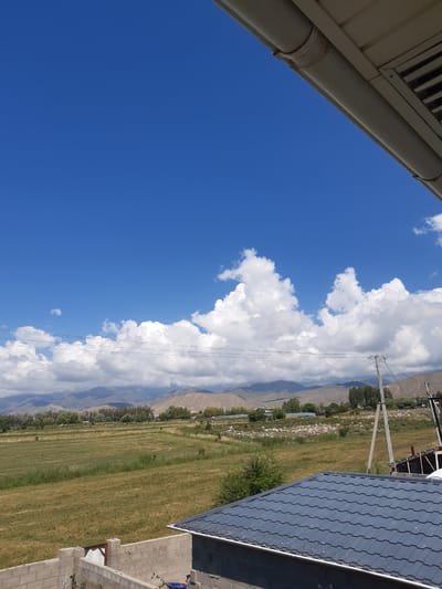 This is a photograph taken from an elevated vantage point, looking out over a rural landscape under a bright blue sky with fluffy white clouds. In the foreground, a gray tiled roof of a building is visible, along with a concrete block wall. Beyond the wall, a vast field of dry grass stretches towards a line of trees and distant mountains. Several utility poles with wires are present in the middle ground, and some scattered buildings can be seen among the trees. The overall impression is one of a sunny day in a somewhat arid, mountainous region, likely in the outskirts of a settlement. The specific location of Progress, Kyrgyzstan, is noted, suggesting this scene depicts a view from or near that area.