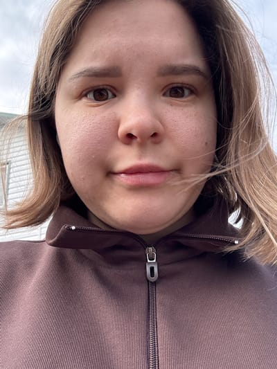 A young woman with brown hair and brown eyes is the focus of the image. She is wearing a brown jacket with a zipper. The background is indistinct but appears to be outside, possibly in the daytime, based on the lighting and a glimpse of a building. The image is a close-up selfie taken at an angle. There is no information in the image to indicate the location is Probuzhdenie, Russia.
