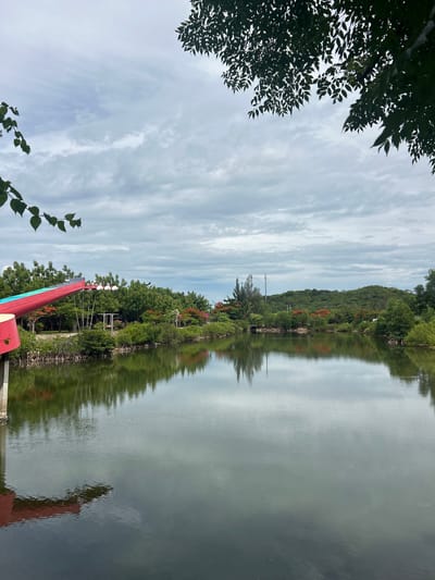 The image depicts a tranquil waterscape in Hoa Thang, Vietnam, characterized by a calm body of water reflecting the overcast sky and lush green vegetation. In the foreground, a red and blue structure, possibly part of a playground or a decorative element, extends into the frame from the left. The water's edge is lined with dense green shrubs and trees, with glimpses of vibrant red flowers interspersed among them, suggesting a tropical or subtropical environment. The background is dominated by rolling hills covered in dense foliage. The sky is filled with dramatic grey clouds, creating a diffused, soft light. The reflection of the trees, sky, and the red structure is clearly visible on the smooth surface of the water. A telecommunications tower is visible in the distance, breaking the natural horizon.