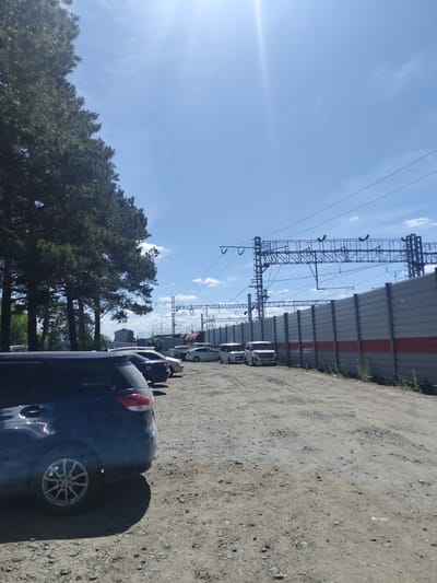 The image depicts an outdoor parking lot situated in the city of Shimanovsk, Russia. In the foreground, the rear left side of a dark blue minivan is visible. Beyond the minivan, several other vehicles are parked in a gravel lot, including a white minivan, a light-colored sedan, and a dark sedan. A tall, gray sound barrier with red stripes runs along the right side of the lot, adjacent to a railway track infrastructure. Overhead power lines and support structures for the railway are prominent features against a bright blue sky with scattered white clouds. To the left, a line of tall, dark green pine trees borders the parking area. The sun is shining brightly, casting shadows on the ground.