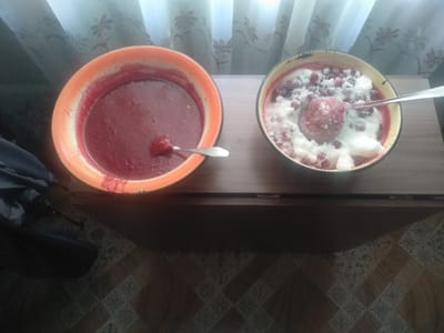 The image shows two bowls of food on a wooden surface, likely a table or counter. The bowl on the left is orange and contains a thick, reddish-purple puree, possibly jam or sauce. A spoon is resting inside this bowl. The bowl on the right is darker colored with a yellowish rim and contains a white, creamy substance mixed with small red berries, possibly cranberries or lingonberries, and is also topped with a scoop of the same creamy substance. A spoon is also present in this bowl. In the background, a light-colored curtain with a floral pattern is visible. To the left of the bowls, a dark-colored bag or backpack is partially visible, resting on a patterned floor. The overall lighting suggests an indoor setting, possibly in a home. The specific location in Osakarovka, Kazakhstan cannot be determined from the visual information provided in the image.