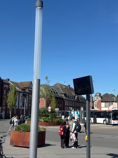 This image captures a vibrant urban street scene in Geldern, Germany, on a clear, sunny day. The sky is a bright, cloudless blue, and strong shadows indicate a high sun.
In the foreground, on the left side of the frame, stands a tall, modern street light or public art installation, featuring a clear, cylindrical tube atop a gray metallic pole. To its right, a black digital display screen, possibly for public transport information, is mounted on another pole.
The mid-ground features a group of approximately three to four people gathered on a paved sidewalk, appearing to be at a bus stop. One individual wears a light-colored jacket with pink and white sections, while another carries a distinctive red bag adorned with a teddy bear or character. Behind them, a blue and white bus is partially visible. Further down the street, two more individuals are walking on the sidewalk, one in a white shirt and backpack.
The background is dominated by a mix of traditional and modern multi-story buildings. Some feature pitched roofs, brick facades, and white window frames, characteristic of German architecture. A white bus with "112" visible on its side is parked or driving on the street in front of these buildings.
Various urban elements are present, including large rectangular planters made of rusty brown metal, filled with green bushes. One planter visibly shows the text "GELD" (likely part of "Geldern" or a related word). Traffic signs include a circular red and white "no entry" sign and multiple yellow "H" signs, indicating bus stops (Haltestelle). A yellow pedestrian signal button is also visible on a pole. A bicycle is partially seen parked at the bottom right of the frame.
