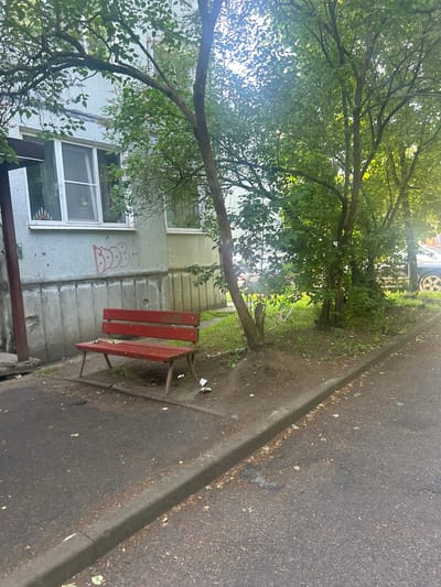 A red, weathered park bench is positioned on a paved area next to a grassy strip and a building in Posyolok Imeni Morozova, Russia. The building, likely an apartment block, has a light-colored tiled facade with a concrete foundation. Red graffiti is visible on the building's facade above the bench. Mature trees with green foliage provide shade, casting shadows on the scene. The paved area appears to be a walkway or parking area and shows signs of wear and scattered debris. In the background, a dark-colored car is partially visible. The lighting suggests it is daytime.