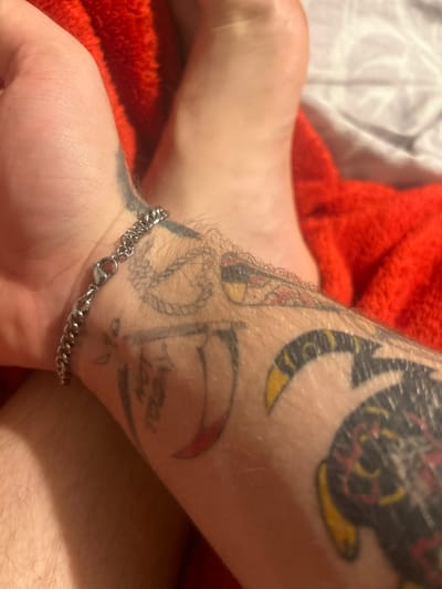 A close-up, overhead shot shows a person's left wrist, adorned with a silver chain bracelet. The wrist is covered in tattoos, including a scythe-like object and text that reads "NOT TODAY". The wrist is resting on a red, textured fabric, possibly a blanket or towel. A portion of a bare foot and leg are visible in the background, also against the red fabric and a white surface. The image does not provide any information about the location within Gelsenkirchen, Germany.