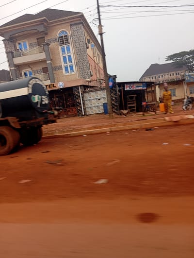 In Imilike, Nigeria, a tanker truck with the partial number "750" visible on its side is parked on a dirt road. The truck's black tank has a white stripe running along its length. To the right of the truck, a three-story building displays the sign "HOTEL" with a circular logo next to it. Further to the right, a smaller structure with a "POS" sign is visible. A person in a yellow patterned outfit stands near this structure, next to a bright orange container. In the background, another building with an arched roof and visible windows is present. A power pole and overhead wires are also in the scene. The ground is covered in red-brown dirt, and the sky is overcast.