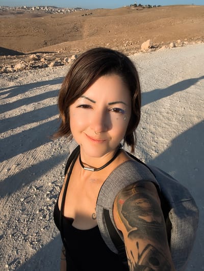 In a selfie taken outdoors, a woman with short brown hair, a nose ring, and subtle face glitter smiles at the camera. She wears a black tank top and a grey backpack. Her left arm is covered in a large, detailed tattoo. The background shows a dry, rocky landscape with a distant city on a hilltop, characteristic of the area surrounding Keidar in the Palestinian Territories. Long shadows stretch across the gravelly ground, indicating it is late afternoon.