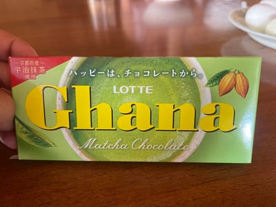 In Pamyati 13 Bortsov, Russia, on a wooden table, sits a box of Lotte Ghana Matcha Chocolate. The box is primarily green with yellow lettering that spells out "GHANA". Below this, in script, it reads "Matcha Chocolate". The top left corner of the box has text in Japanese: "京都産" (Kyoto product) and "宇治抹茶" (Uji matcha), with "使用" (used) below it. To the right of the main text, there are two cocoa pods and a green leaf illustration. The image is a close-up shot, with a hand holding the left side of the box, obscuring part of the text. In the blurred background, a white plate with what appears to be hard-boiled eggs is visible.