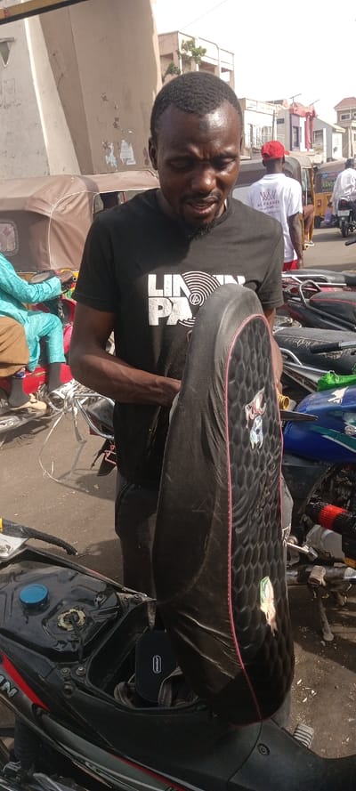 A dark-skinned adult male is positioned slightly right of center, standing on an asphalt street in Fagge D2, Nigeria. He wears a black short-sleeved t-shirt with a white 'LINKIN PARK' graphic and dark trousers. His face and forehead exhibit perspiration. He is actively holding a black motorcycle seat upright; the seat features a textured, hexagonal quilted pattern, a prominent red stitched border, and several cartoon-style decals. This seat is detached from a black motorcycle, partially visible in the lower-left foreground, which has red side accents and a blue fuel cap. The motorcycle's under-seat storage compartment is open, containing a black rectangular case labeled 'PowerBanker' and a piece of folded fabric. In the mid-ground and background, other individuals are visible amidst various vehicles. One person, wearing a light-blue patterned garment, is seated on a red auto-rickshaw (Keke Napep). Another individual wears a white t-shirt with 'AL FAKHER' in white text and a red baseball cap, standing near another auto-rickshaw. Multiple other auto-rickshaws and motorcycles are parked or in transit. Light-colored, multi-story buildings line the background under a clear, bright sky, with one wall showing areas of peeling posters.