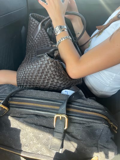 The image depicts a close-up, downward-angled view of a person seated within a vehicle. The visible portion includes a tanned right arm, a right hand with red-painted fingernails holding an unseen object, and part of a torso wearing a white ribbed tank top. The right wrist is adorned with a silver-toned rectangular-faced watch, a thin silver bangle, and a thick silver Cuban link chain bracelet.
A large, dark brown, woven leather tote bag is positioned on or near the person's lap. Beneath this bag, a large, dark-colored duffel bag is visible, featuring a subtle monogram pattern, two prominent horizontal gold-toned zippers, and black leather straps with gold-toned buckles. A white tag with a blue arrow pointing left is partially visible between the two bags. Sunlight casts distinct shadows across the lower duffel bag, indicating an external light source. The immediate environment consists of dark, indistinct vehicle interior elements. The scene is consistent with travel, potentially occurring in Parikia, Greece, although no specific landmarks or external views confirming this location are present within the image.