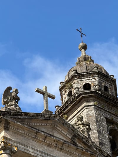 A low-angle, close-up shot captures the upper architectural elements of the Cathedral of Santiago de Compostela in Spain, set against a vibrant, partially clouded blue sky. The focus is on the weathered stone facade, featuring intricate carvings and details. On the left, a stone statue of an angel with outstretched wings stands atop the building. To its right, a large stone cross is prominently displayed. Further to the right, a domed bell tower, adorned with smaller decorative elements and patches of greenery, rises towards the sky, topped by a smaller, ornate metal cross. The stone surfaces show signs of age, with moss and lichen growing in crevices, lending a sense of historical depth to the structure. The overall impression is one of architectural grandeur and timelessness.