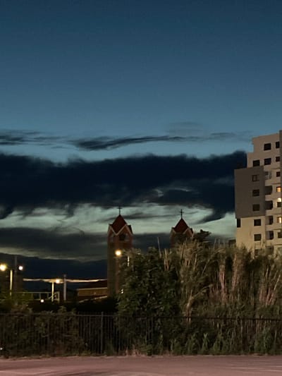 The image depicts an urban or suburban landscape in Bjeliši, Montenegro, captured at twilight or early night. The upper sky is a dark, deep blue, transitioning into a horizontal band of dense, dark clouds that extend across the mid-section of the frame. Beneath this dark cloud layer, lighter, more textured clouds are visible.
Two prominent structures, resembling church towers, stand in the mid-ground. Both towers feature distinctive red, gabled roofs topped with crosses. Their facades appear to be of a light masonry material, with arched openings, some of which are internally illuminated. To the right of these towers, a multi-story building of light-colored, possibly beige, construction rises. This building exhibits numerous dark, rectangular windows, with several windows on different floors showing internal illumination. Its upper section features a stepped or terraced design.
In the mid-ground, a dense line of tall, dry reeds and dark green foliage runs horizontally, partially obscuring the lower portions of the visible structures. A dark metal fence, likely a security barrier, is positioned in front of this vegetation. In the lower-left, multiple streetlights are visible, casting light onto the scene. The foreground consists of a reddish-brown surface, consistent with a road or pavement. No individuals or specific actions are discernible within the frame.