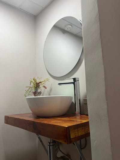 The image shows a bathroom sink and vanity in a bathroom located in Las Palmas de Gran Canaria, Spain. The sink is white porcelain and is mounted on a wooden vanity. The vanity has a rustic, distressed wood finish and is supported by a chrome metal frame. There is a chrome faucet mounted on the counter. There is a round mirror above the sink. There are plants in a glass vase on the counter. The walls of the bathroom are painted white. The floor is not visible in the image. The bathroom is lit by a single overhead light fixture. The bathroom appears to be clean and well-maintained.