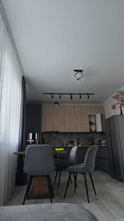 This image depicts an indoor scene, likely a kitchen or dining area, in Bulgakovo, Russia. The room features modern, minimalist furniture and a neutral color palette.
The primary subjects are several chairs with grey upholstery and black metal legs, arranged around a counter or table area. Behind the chairs, there are kitchen cabinets with a wood-grain finish on the upper cabinets and dark grey on the lower ones. A dark grey countertop or backsplash is visible, with various items, possibly kitchen supplies or decor, placed on it. To the left of the cabinets, there is a wall with a black light fixture holding three glass shades. Further to the left, a set of black rods with several bottle-shaped holders is mounted on the wall, suggesting a wine rack. The floor appears to be wood-look tile. A large window with white blinds is visible in the foreground, on the left side of the image. The lighting suggests it is daytime, with artificial light from the fixtures. There are no people visible in the image.