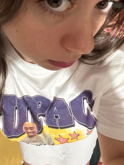 The image is a close-up, overhead shot of a young woman wearing a white t-shirt with a graphic print. The t-shirt features large purple lettering, partially visible, and a smaller image of a man's face, possibly Tupac Shakur, on a yellow background with red stars. The woman's face is visible from the nose up, with her brown eyes looking slightly off-camera and her lips closed. Her dark brown hair is partially visible. The background is blurred and appears to be a solid yellow surface. There is no discernible location in the city of Garmen, Bulgaria.