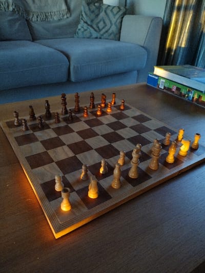 The image displays an indoor scene, consistent with a living room, located in New Forest, United Kingdom. A wooden chessboard, featuring alternating light and dark squares, is centrally positioned on a dark wooden coffee table. Light-colored (pale wood) and dark-colored (stained wood) chess pieces are meticulously arranged on the board, appearing to be in their initial setup for a game. A strong, warm, golden light source, originating from the left (viewer's perspective), illuminates the lower left edge of the chessboard and causes the light-colored chess pieces to glow distinctly. In the background, a light grey fabric sofa is visible, accented by a plain light grey fringed throw draped over its left side and a geometrically patterned cushion on its right. To the right of the sofa, a window covered by vertical striped curtains is partially visible, also catching streaks of the golden light. A stack of two books, including one with 'Ravensburger' visible on its spine or cover, rests on the coffee table beside the curtains. The scene is otherwise dimly lit, characterized by the directional golden light creating prominent highlights and shadows. No individuals are present.