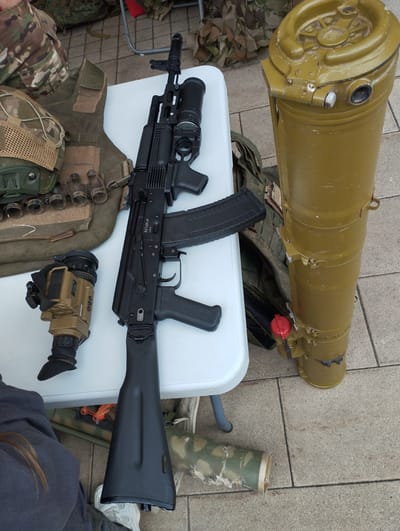 A flat lay image displays military equipment on a white table and the surrounding tiled ground. The central object is a black assault rifle with a magazine inserted and an optic mounted on top. To the left of the rifle, on the table, is a tan monocular or night vision device. Behind the rifle and to the left is a piece of body armor with several pouches and a helmet with a mesh covering.

To the right of the rifle, on the ground, stands a large, olive-drab cylindrical object, possibly a missile or rocket launcher casing, with a red cap on a small component attached to its side. Further to the right and partially visible on the ground is a camouflage-patterned fabric, possibly part of a backpack or camouflage netting. A person's legs, wearing dark pants and white sneakers, are visible at the bottom left of the image, indicating a person is present, though not fully in frame.

The scene appears to be set outdoors, given the tiled ground resembling a patio or walkway. The lighting suggests it could be daytime, but the weather is not discernible. The overall impression is a display or staging of military or tactical gear.

Visible text includes "AK74M-M" and "545x39" on the rifle. The cylindrical object has some markings, including "609-04" and numbers that are partially obscured.