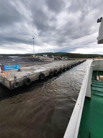 The image shows a dock at Terminal 2 in Shcheleiki, Russia. The dock is made of concrete and is lined with tires to protect ships. There is a ramp leading from the dock to the shore, where piles of dark material are visible. The sky is overcast with heavy clouds. Part of a green-decked vessel is in the foreground, on the right side of the image.