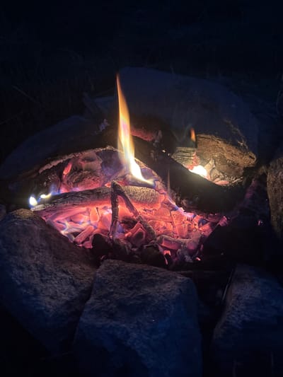 The media file is a close-up, high-angle shot of a campfire at dusk or night. The main subjects are the burning embers and logs within a ring of rocks. The fire is predominantly orange and red, with hints of blue and purple in the hotter coals. A single, bright flame extends from the left side of the frame, casting a warm glow on the surrounding rocks and some charred wood. The texture of the rough, grey rocks contrasts with the soft, glowing embers. The background is dark and out of focus, suggesting a natural setting like a campsite or wilderness area. There are no people visible, and the scene evokes a sense of warmth and solitude. The time of day is clearly nighttime or late dusk due to the darkness of the background and the prominence of the firelight. There is no visible text in the media file. The location context is Batak, Bulgaria.