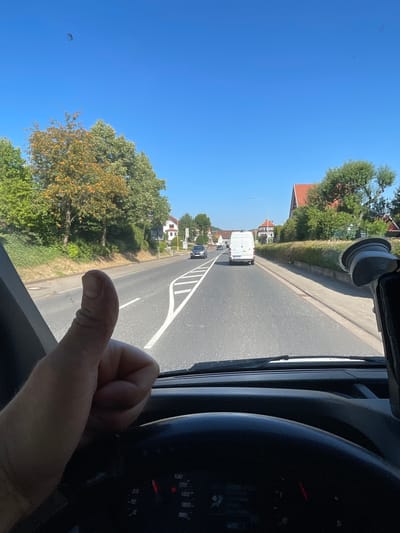 An outdoor, eye-level, forward-facing shot captures a driver's hand giving a thumbs-up from inside a vehicle. The driver's thumb is prominently positioned in the foreground, and a portion of the steering wheel and dashboard gauges are visible at the bottom of the frame. The vehicle is on a paved road in what appears to be a suburban area. A white cargo van is driving ahead on the road, followed by a dark-colored car. The road is lined with trees and houses with red-tiled roofs on either side. The sky is clear and blue, indicating good weather. The scene is likely in Wartenberg, Germany, given the architecture and the style of the vehicles.