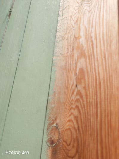 The image is a close-up, low-angle shot of a wooden surface that is half painted a light green and half unpainted wood with a visible grain pattern. The painted section shows several small imperfections, possibly from the paint application or the wood surface itself. The unpainted wood has prominent circular knots and vertical grain lines. The lighting suggests an outdoor environment, possibly a wall or fence. The lower-left corner of the image displays the text "HONOR 400" in white. This scene is located in the city of Эрхирик, Russia, and it depicts an architectural detail of a wooden structure. There are no people or animals present in the frame.