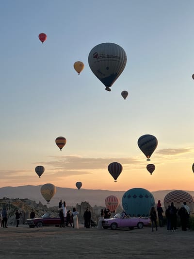 The image, presented in a vertical orientation, captures a stunning hot air balloon display at what appears to be sunrise over the distinctive landscape of Göreme, Turkey. The sky transitions from a soft, warm golden hue near the right horizon to a lighter blue towards the left, indicating either dawn or dusk.

Numerous hot air balloons, varying in size, color, and pattern, are visible across the expansive sky. A prominent large white balloon, positioned in the mid-ground, features "Kaya Balloons" and "www.kapadokyayaballoons.com" printed on its side. Other balloons include a large teal-colored one with geometric patterns, a red-and-white striped balloon with "Unitemp" text, and many smaller, colorful balloons with stripes, checks, and solid colors ascending and floating in the distance. Some balloons appear to be still very low, possibly preparing for flight, while others are already high above.

On the right side of the frame, a crowd of people is gathered on a dusty, rocky terrain, observing the spectacle. Several individuals are standing near a striking vintage pink or purple convertible car parked on the ground. In the background, the unique undulating rock formations and "fairy chimneys" characteristic of Cappadocia are visible under the warm light. The scene conveys the popular and breathtaking event of hot air ballooning in a picturesque setting.