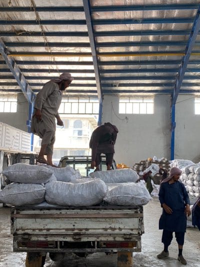 The photo depicts an interior warehouse scene in Shurandam, Afghanistan. Three men are visible: two on the back of a small flatbed truck loaded with large white sacks, presumably produce, and one standing on the ground near the back of the truck. The two men on the truck appear to be loading or arranging the sacks. The man on the ground is wearing a blue robe, while those on the truck wear what appears to be traditional clothing. More stacked sacks can be seen in the background. The warehouse has a high ceiling with metal supports and a roof, illuminated by natural light filtering through windows. The overall impression is that of a warehouse or storage facility related to agriculture or commerce.